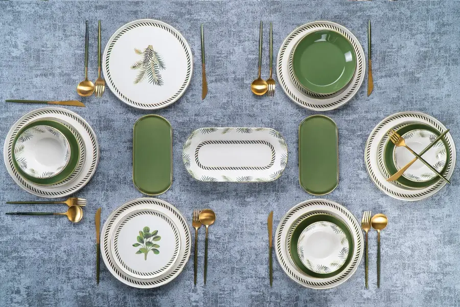 Elegant table setting featuring green and white botanical dinnerware with rope-textured borders, including dinner plates, bowls, and serving dishes decorated with fern and leaf motifs, arranged with gold flatware on a blue textured surface